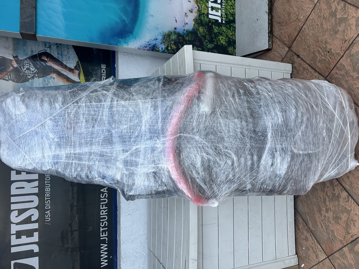 Nose of a board bag wrapped tightly in stretch wrap, with a foam pool-noodle rail visible underneath at the edge